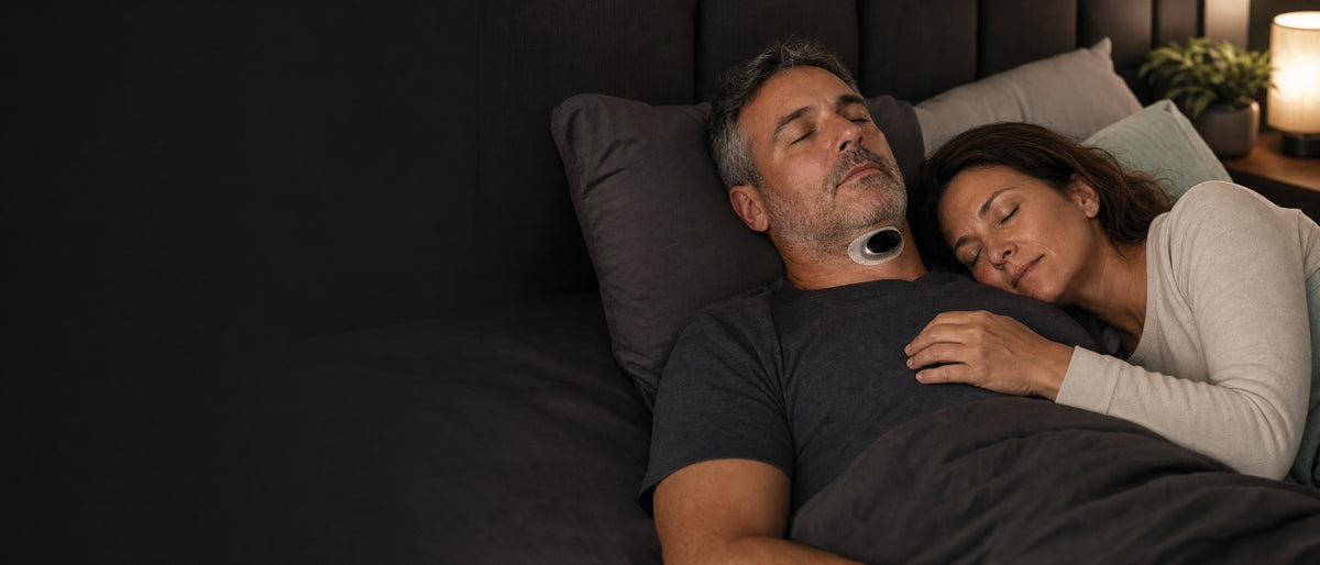 A couple asleep in a dimly lit bedroom, the man wearing the VoroHal Sleep Aid under his chin